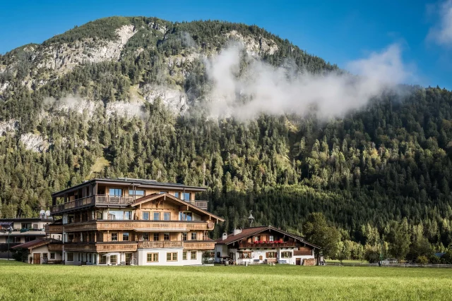 Alpenhaus vor bewaldetem Berg mit Wolken.