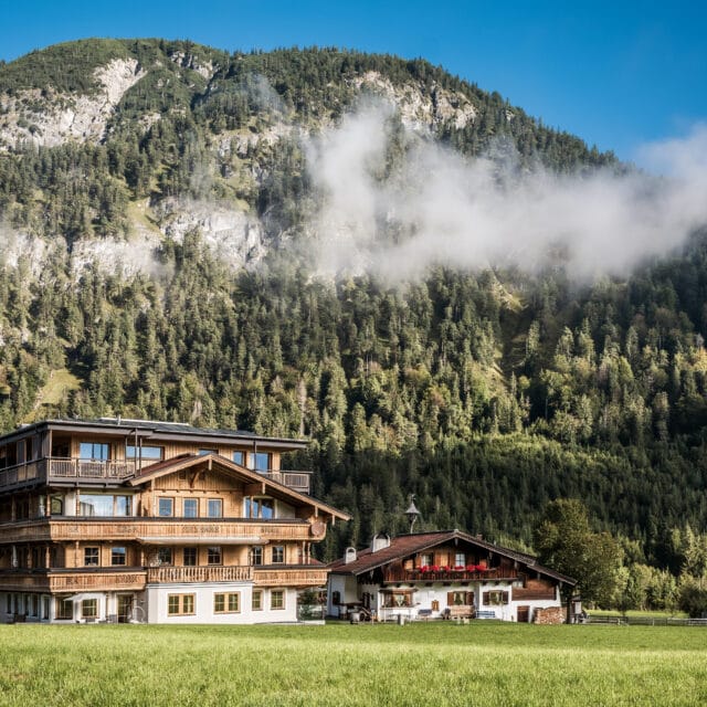 Alpenhaus vor bewaldetem Berg mit Wolken.